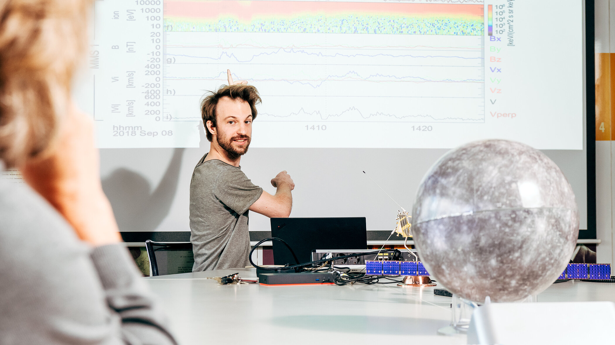 A man is smiling as he presents scientific charts on a large screen. On the table in front of him is a large celestial globe. A person in the foreground is watching him.