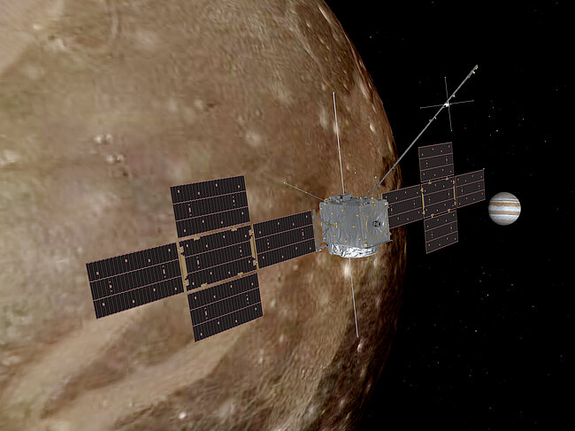 An artist's rendering of the ESA JUICE spacecraft in space, with its solar panels extended wide in a cross-shaped formation. It is flying close to Jupiter's massive, crater-covered moon Ganymede, while the striped gas giant Jupiter can be seen in the background.