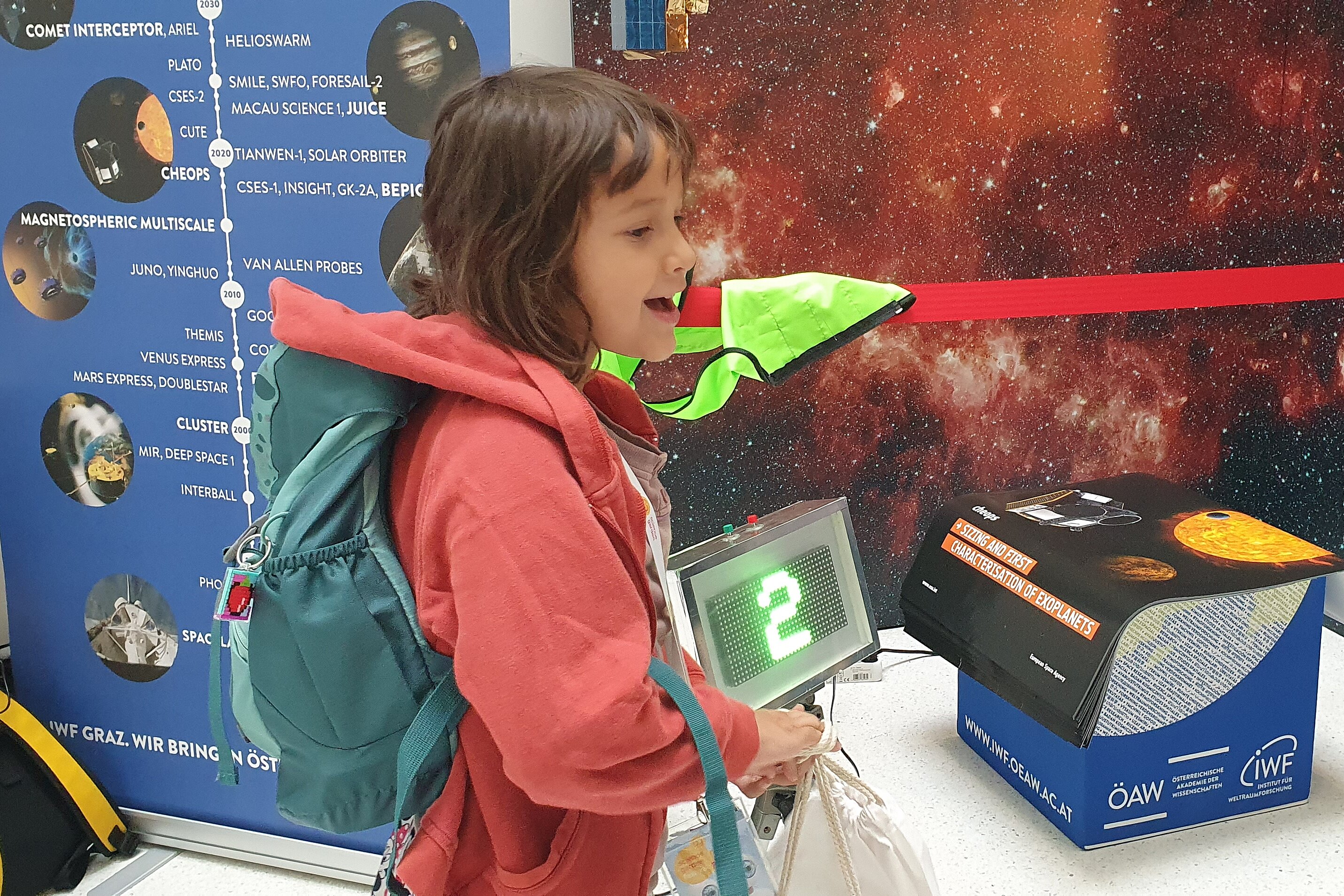 A smiling child wearing a red jacket is standing in front of a museum exhibit. In the background, there is a blue information board featuring space missions and a photo wall depicting star galaxies.