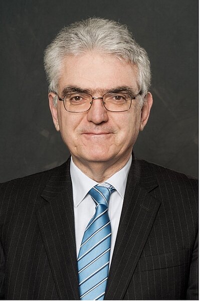 Oberkörper eines älteren Mannes mit grauen Haaren in einem dunklen Nadelstreifenanzug, weißem Hemd und hellblau gestreifter Krawatte vor dunklem Hintergrund.