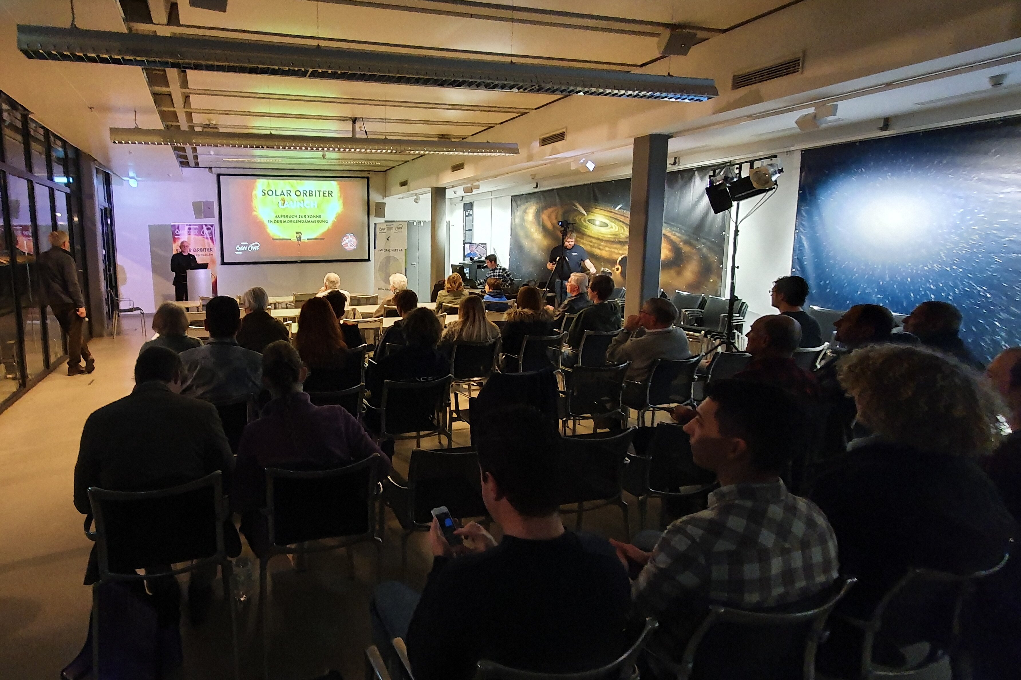 An audience sits in a darkened lecture hall, looking at a screen projection titled "SOLAR ORBITER". On the left, a person stands at a lectern against a brightly lit background, while chairs are arranged in rows throughout the hall.