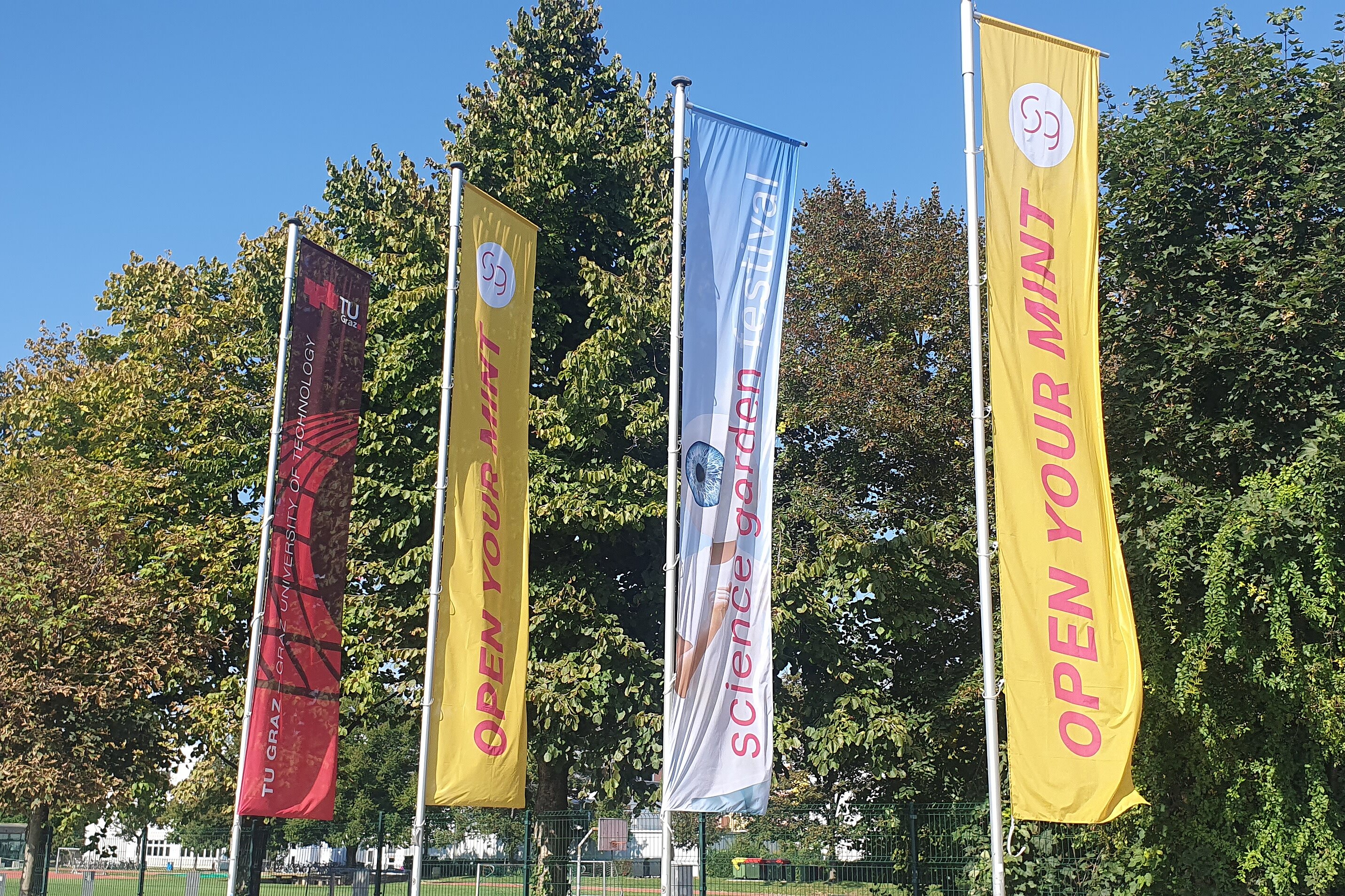 The picture shows four banners with the text "TU Graz", "Open your mint" and "Science garden festival".  In the background, there are trees and a sports field.