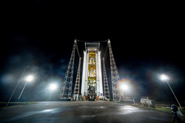 A photo showing a rocket launch pad at night. At the centre of the image is a tall rectangular cream-coloured building, with a thin white rocket inside. Platforms at various heights give people access to different parts of the rocket. The building is surrounded by metal pylons.