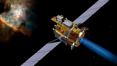 A golden, cylindrical satellite with two long solar panel wings emits a bright blue ion beam. In the dark expanse of space in the background, a colorful, red-orange glowing gas nebula is visible in the upper left corner.