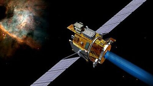 A golden, cylindrical satellite with two long solar panel wings emits a bright blue ion beam. In the dark expanse of space in the background, a colorful, red-orange glowing gas nebula is visible in the upper left corner.