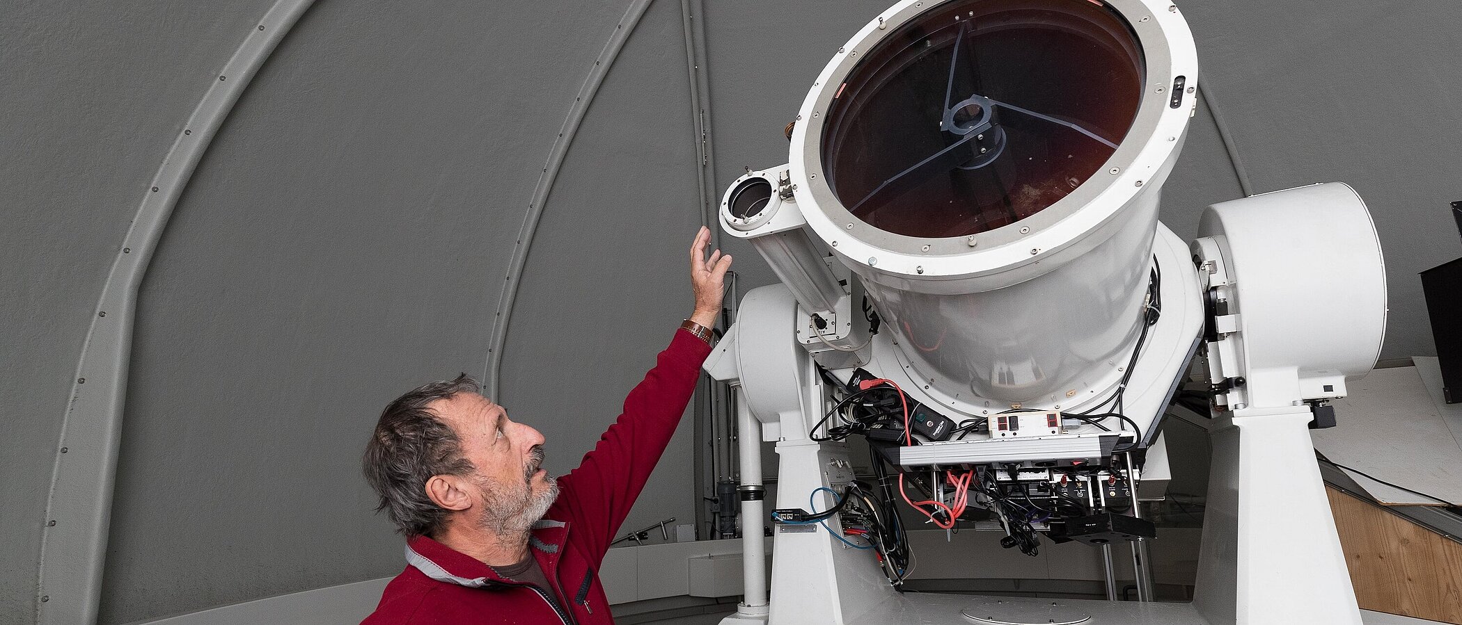 A bearded man in a red sweater looks up and reaches out his hand toward a large, white reflecting telescope. The telescope is mounted inside a round, gray observatory dome.