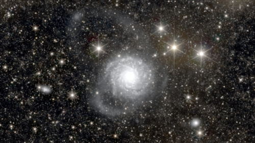 A circular telescopic image shows a pitch-black universe dotted with countless small, brightly shining stars. At the center of the view is a nebulous, pale blue celestial body with a particularly bright center.