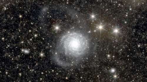 A circular telescopic image shows a pitch-black universe dotted with countless small, brightly shining stars. At the center of the view is a nebulous, pale blue celestial body with a particularly bright center.