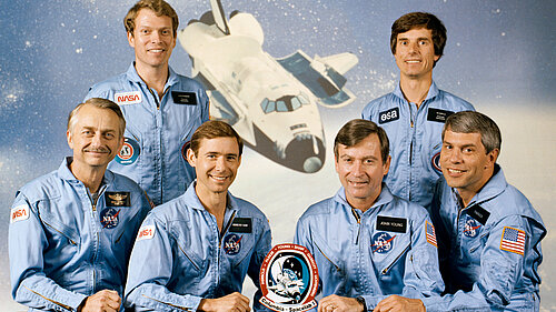 Six smiling astronauts in light blue jumpsuits are posing together behind a table against a space backdrop. In the center behind them is an illustration of the Space Shuttle Columbia landing.