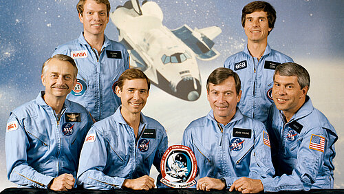 Six smiling astronauts in light blue jumpsuits are posing together behind a table against a space backdrop. In the center behind them is an illustration of the Space Shuttle Columbia landing.