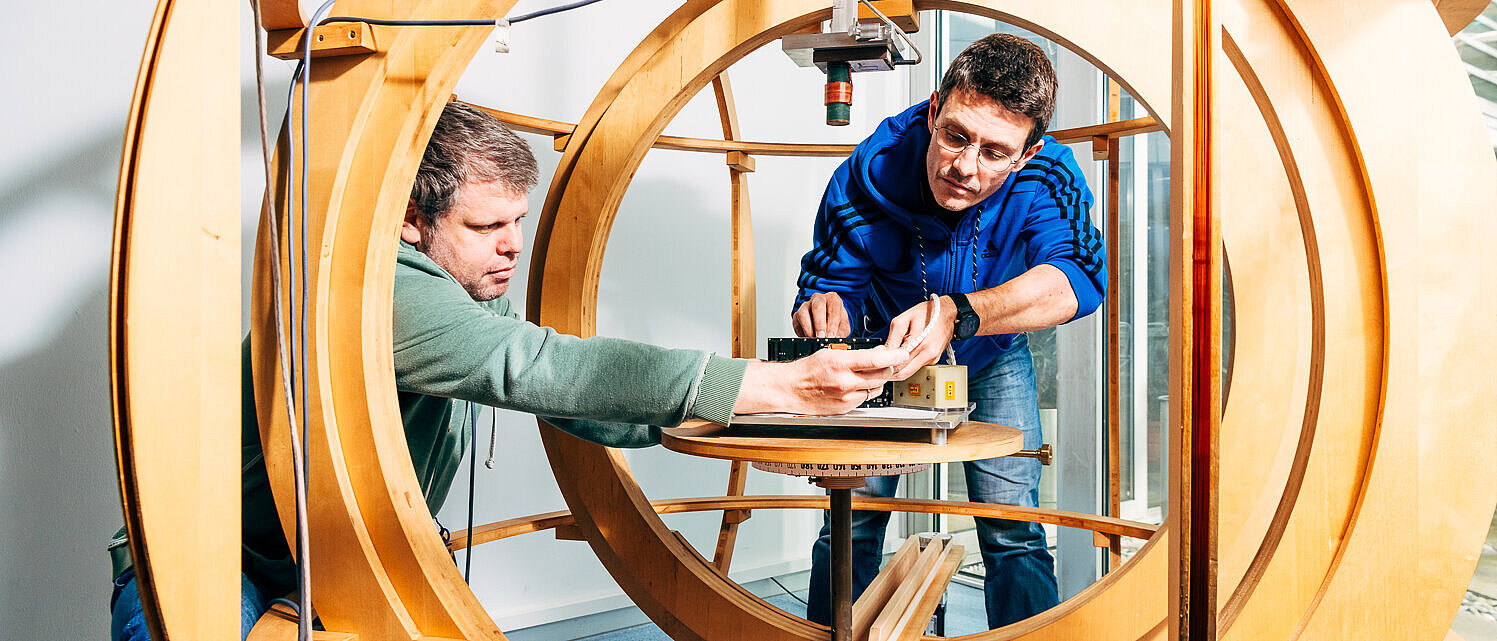 Two men are working on a laboratory device made of large, nested wooden rings. They are both focused on adjusting the settings on a platform in the center of the device.