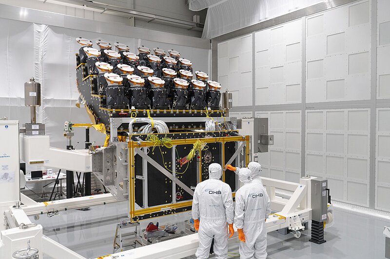 In the photo three people in white overalls and caps stand in front of the instrumentation in a white hall. The equipment, about twice as tall as the people in the picture, consists of a black four-stepped-platform that supports 24 cameras. These resemble cylinders flaring at the top, not unlike big ice-cream cones. Each cylinder is wrapped in black material; the top flare has a metallic finish and a white lid.