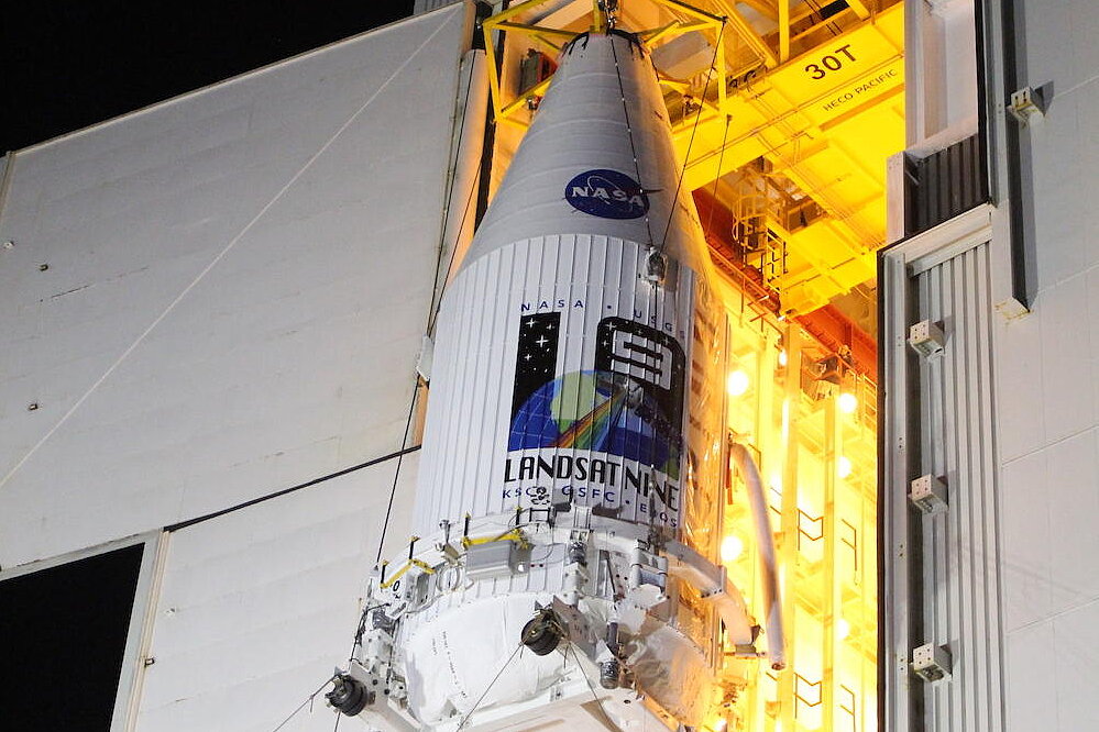 A nighttime photo shows the white payload fairing of a launch vehicle being hoisted by cranes in an assembly hall. The blue NASA logo and the colorful “LANDSAT NINE” mission emblem are clearly visible on the fairing.