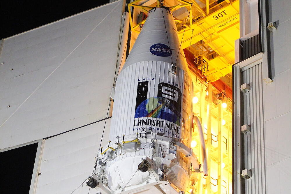 A nighttime photo shows the white payload fairing of a launch vehicle being hoisted by cranes in an assembly hall. The blue NASA logo and the colorful “LANDSAT NINE” mission emblem are clearly visible on the fairing.