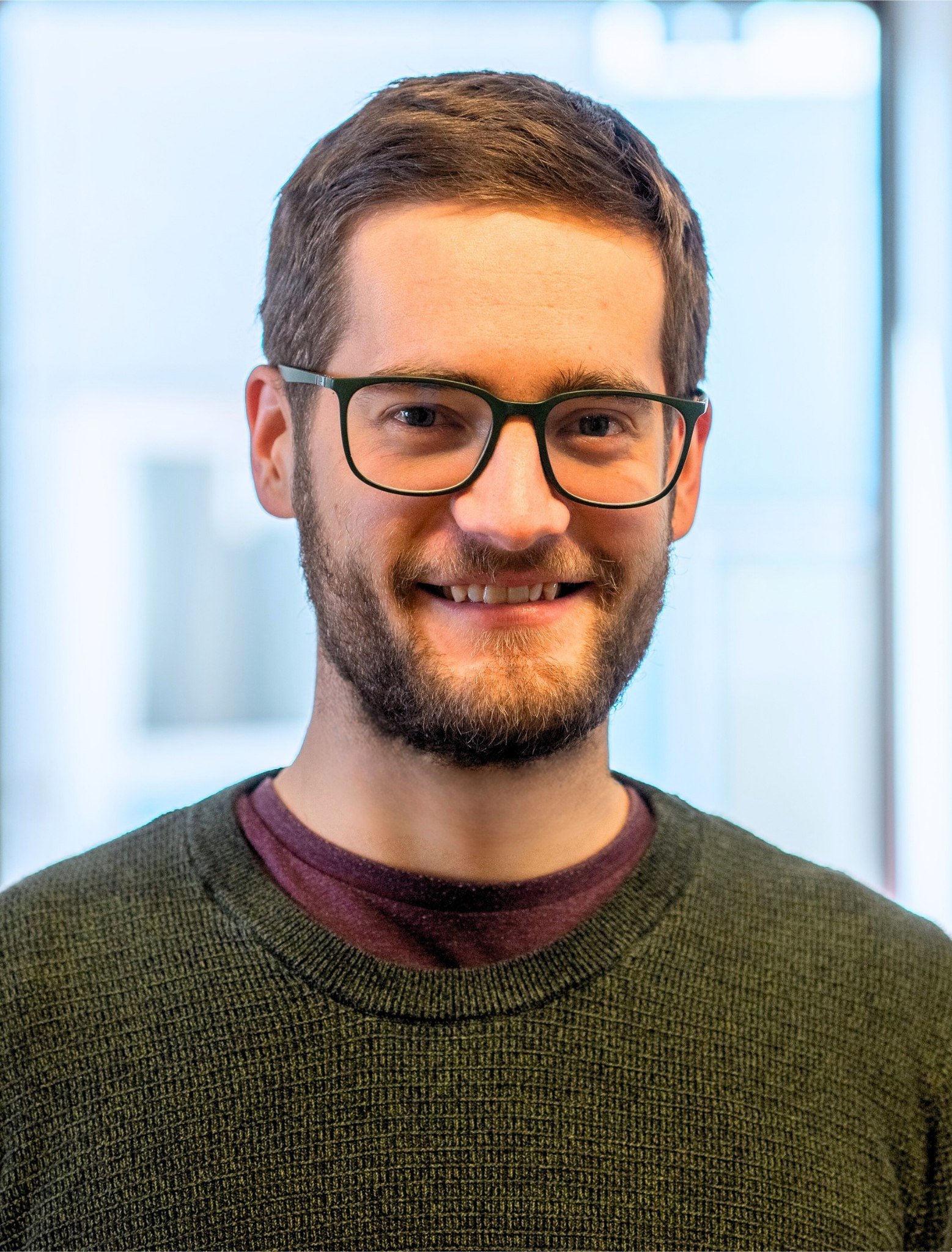 Oberkörper eines Mannes mit kurzem braunem Haar, Brille und grünem Pullover vor unscharfem Hintergrund.